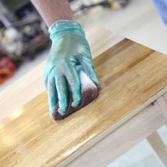 Man's hand wearing blue gloves while using a sponge staining pad on a wooden surface.