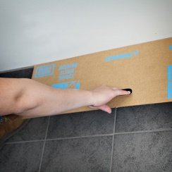 Man's hand holding a cardboard paint spray shield at the junction of a wall with the floor